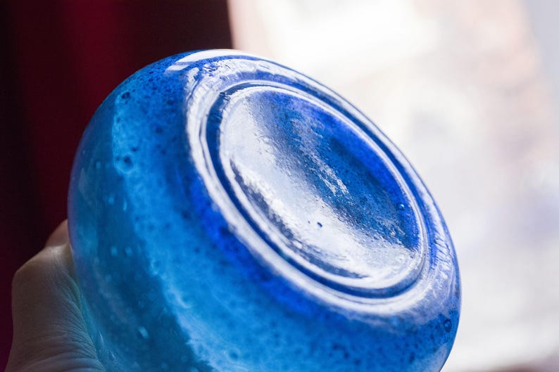 May include: Close-up of a blue glass bottle base. The glass has a textured surface with a glossy finish. The bottle is a vibrant shade of blue, with a circular indentation in the centre.