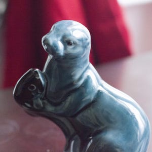 Puede incluir: Una figurita de cerámica azul de una nutria sosteniendo un pez en sus patas. La nutria está de pie sobre una superficie roja.