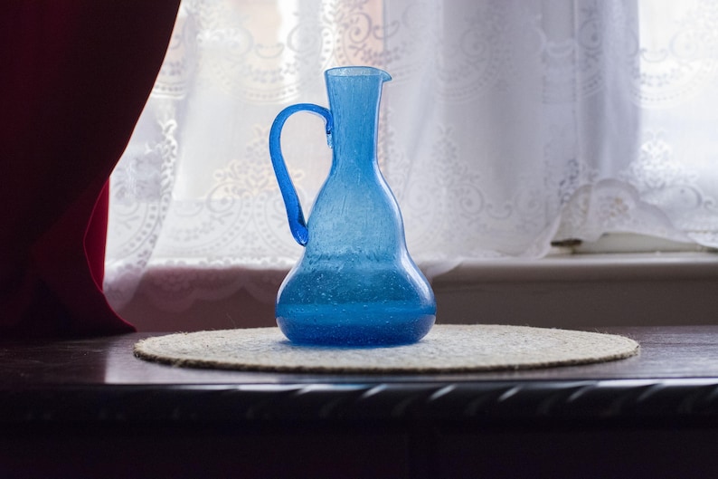 May include: A blue glass jug with a handle, placed on a round, woven placemat. The jug has a rounded base and a narrow neck with a spout. The background features a window with white curtains and a dark wooden surface.