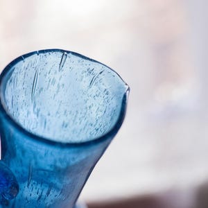 May include: Close-up of a blue glass jug with a handle. The jug has a wide opening and a slightly textured surface. The glass is a vibrant shade of blue, and the background is blurred.