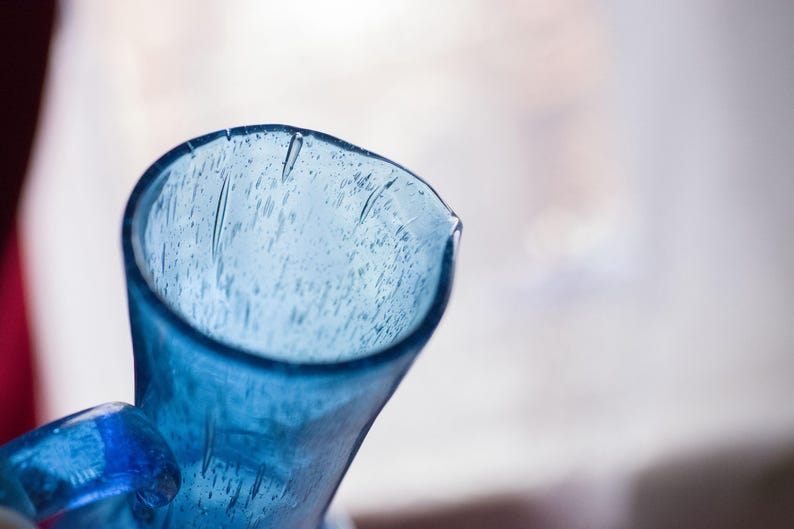 May include: Close-up of a blue glass jug with a handle. The jug has a wide opening and a slightly textured surface. The glass is a vibrant shade of blue, and the background is blurred.