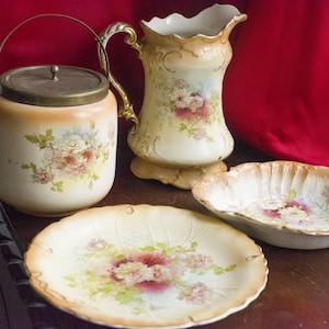 Puede incluir: Conjunto de piezas de servicio de porcelana antiguas, que incluyen un tarro con tapa, una jarra, un plato y una fuente. Cada pieza está decorada con diseños florales en tonos rosa, blanco y verde sobre un fondo crema.
