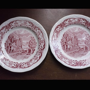 May include: Two white ceramic plates with a red and white landscape design. The plates have a decorative border and a central image of a town scene. The plates are on a dark brown surface.