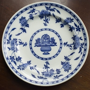 May include: A white ceramic plate with a blue and white floral design. The plate features a central design of a basket with flowers and a border of repeating floral motifs.