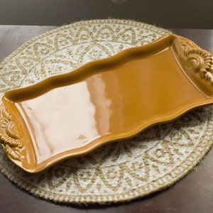 Puede incluir: Una bandeja rectangular de cerámica marrón con un borde ondulado y diseños florales decorativos. La bandeja está sobre un mantel tejido blanco y marrón.