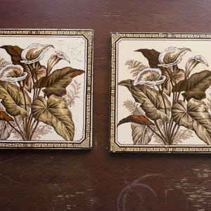 Può includere: Due piastrelle decorative quadrate con un motivo floreale. Ogni piastrella presenta uno sfondo color crema con un'illustrazione dettagliata di calle e grandi foglie nei toni del marrone e del beige. Le piastrelle hanno un bordo decorativo.