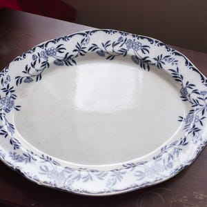 Puede incluir: Plato ovalado de cerámica blanca con un borde floral azul. El plato tiene una forma ligeramente irregular y un acabado brillante. El diseño floral incluye hojas y flores. El plato está sobre una superficie marrón oscuro.