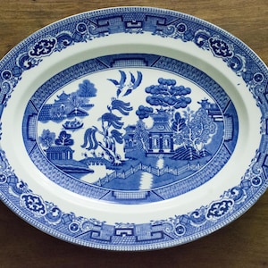 May include: An oval ceramic platter featuring a blue and white willow pattern. The central design depicts a landscape with a pagoda, a bridge, a willow tree, and two birds in flight. The rim is decorated with a geometric and floral border pattern.