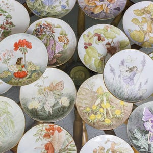 Pode incluir: Uma coleção de 12 pratos decorativos com ilustrações de fadas caprichosas. Cada prato tem um design diferente com fadas, flores e cenas da natureza. Os pratos são brancos com bordas douradas e são perfeitos para exibir ou colecionar.
