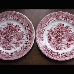 May include: Two matching antique plates with a red and white transferware design. The plates feature a rural landscape scene with trees, a house, and a person walking on a path. The rims have a floral pattern.