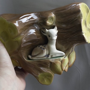 Puede incluir: Una maceta de cerámica con forma de tronco con una figura de cervatillo en su interior. La maceta es marrón con detalles verdes y amarillos, y el cervatillo es blanco con manchas marrones. Las hojas son verdes y amarillas.
