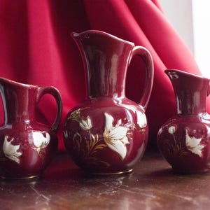 May include: Three maroon ceramic jugs with white floral designs and gold accents. The jugs vary in size, with the largest in the centre. They are set against a red backdrop.