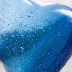 May include: Close-up of a blue glass vase with a rounded shape. The vase has a glossy finish and is covered in small water droplets. The colour transitions from a lighter blue to a deeper shade.