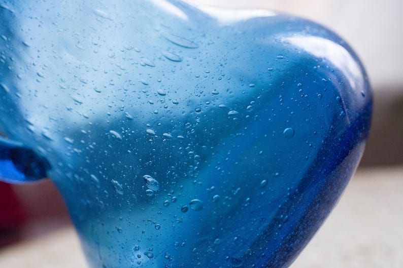 May include: Close-up of a blue glass vase with a rounded shape. The vase has a glossy finish and is covered in small water droplets. The colour transitions from a lighter blue to a deeper shade.