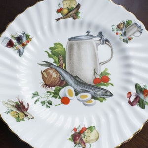 May include: A white ceramic plate with a gold rim features a hand-painted design of a fish, vegetables, eggs, and a beer stein. The plate is perfect for serving a variety of dishes.