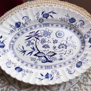 Puede incluir: Una fuente ovalada de cerámica blanca con borde festoneado. La fuente está decorada con un patrón floral azul. El diseño incluye flores estilizadas, hojas y otros elementos botánicos. La fuente está colocada sobre un salvamanteles beige tejido.
