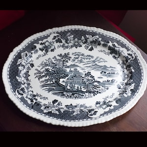 May include: An oval serving platter with a blue and white Chinoiserie design. The platter features a detailed landscape scene in the centre, surrounded by a floral border and a patterned edge. The rim has a scalloped design.