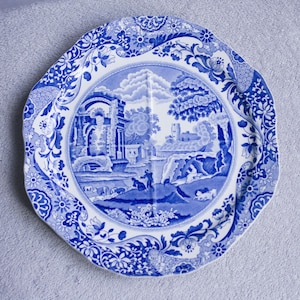 Könnte beinhalten: Blau-weiße Zierplatte mit gewelltem Rand. Die Platte zeigt eine zentrale Landschaftsszene mit Gebäuden, Bäumen und Figuren, umgeben von einem floralen und scrollartigen Rand. Hergestellt aus Keramik.