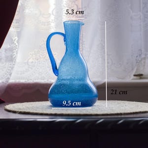 May include: A blue glass jug with a handle, 21 cm tall, 9.5 cm wide, and 5.3 cm across the top. The jug is on a round, beige mat. The background includes a window with white curtains and a dark wooden surface.