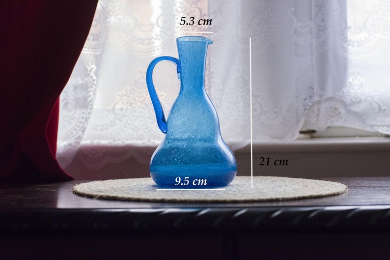 May include: A blue glass jug with a handle, 21 cm tall, 9.5 cm wide, and 5.3 cm across the top. The jug is on a round, beige mat. The background includes a window with white curtains and a dark wooden surface.