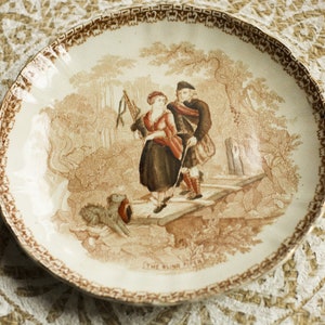 Puede incluir: Un plato de cerámica vintage con un diseño marrón y blanco. El plato presenta una escena de dos personas en un entorno boscoso. El texto "The Blind Piper" está impreso en la parte inferior del plato.