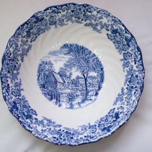 May include: A white and blue ceramic bowl with a detailed landscape scene in the center. The bowl features a blue floral pattern around the rim and a scalloped edge. The central image depicts a water mill and trees.