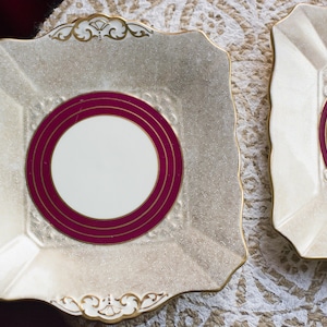 Puede incluir: Dos platos cuadrados de color crema con superficie texturizada y ribete dorado. Cada plato presenta un centro blanco rodeado por dos anillos burdeos. Los platos tienen detalles dorados decorativos en los bordes.