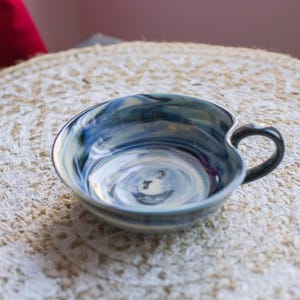 May include: A blue and white ceramic heart-shaped bowl with a handle. The bowl has a swirl pattern and is glazed in a light blue colour.