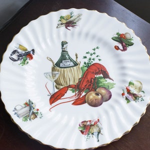 May include: White ceramic plate with a gold rim featuring a lobster, a bottle of wine, and various other food items. The plate is decorated with a white and green floral design.