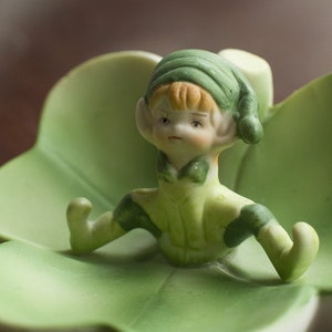 Puede incluir: Una figurita de cerámica verde de un duende sentado en un plato con forma de trébol. El duende lleva un sombrero verde y un atuendo verde y blanco.