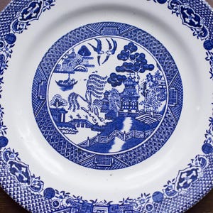 Może przedstawiać: Biały talerz ceramiczny z niebiesko-białym wzorem przedstawiającym chiński krajobraz. Talerz ma podwyższony rant i gładką powierzchnię.