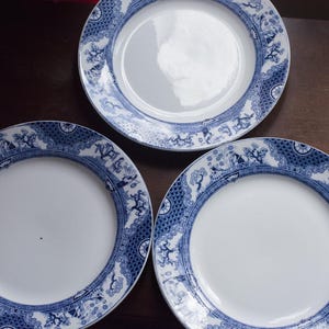 Peut inclure: Trois assiettes en céramique blanche avec une bordure décorative bleue. La bordure présente un motif détaillé d'arbres, de personnages et de formes géométriques. Les assiettes sont disposées sur une surface marron foncé.