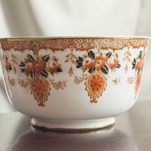 Può includere: Una tazza da tè in porcellana bianca con un bordo dorato e un intricato motivo floreale in tonalità arancio, marrone e nero.
