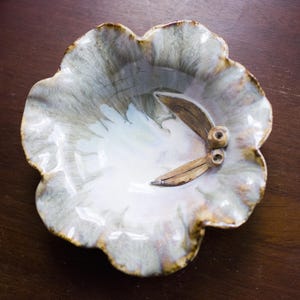 Puede incluir: Un plato de cerámica hecho a mano con una forma orgánica única. El plato presenta un interior blanco con detalles grises y marrones, y un elemento decorativo que se asemeja a vainas de semillas. El borde tiene un borde marrón.