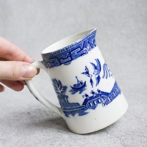 Puede incluir: Jarra de cerámica blanca con un diseño azul de patrón Willow. La jarra tiene un asa y un borde acampanado. El diseño azul y blanco representa un paisaje escénico con pájaros, un barco y un puente. La jarra se sostiene en ángulo.
