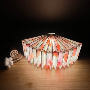May include: A white and pink paper lampshade with a fan-like design. The lampshade is lit from within, casting a warm glow.
