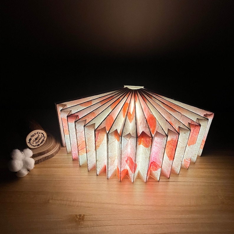 May include: A white and pink paper lampshade with a fan-like design. The lampshade is lit from within, casting a warm glow.
