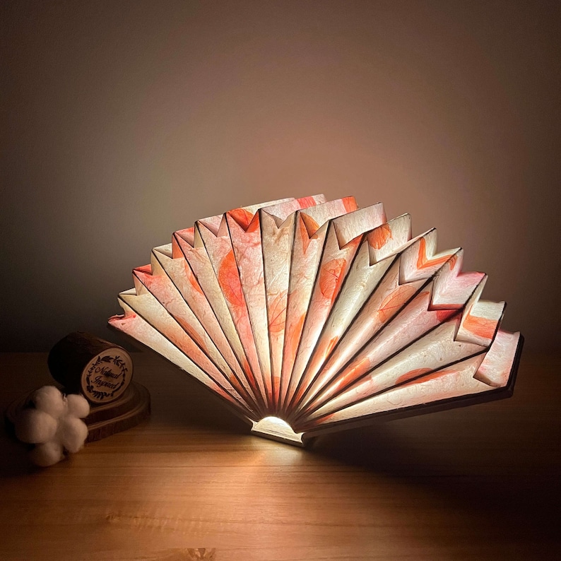 May include: A decorative lamp shaped like a fan, made of paper with a pink and orange floral pattern. The lamp is lit and casting a warm glow.