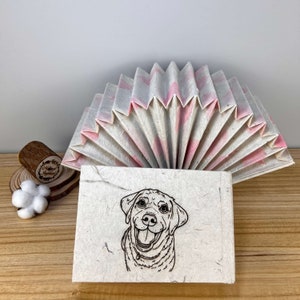 Puede incluir: Un diario de papel blanco con un dise&ntilde;o de abanico de papel rosa y blanco en la portada. El diario tiene una ilustraci&oacute;n en blanco y negro de un perro en la parte delantera.