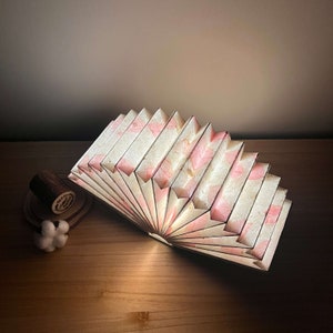Puede incluir: Una pantalla de l&aacute;mpara de papel blanco y rosa con un dise&ntilde;o en forma de abanico. La pantalla est&aacute; hecha de papel fino y en capas y tiene un aspecto delicado y femenino.