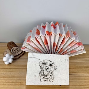 May include: A handmade paper book with a fan-shaped cover featuring orange leaf designs. The book is open to reveal a black and white illustration of a dog with a happy expression.