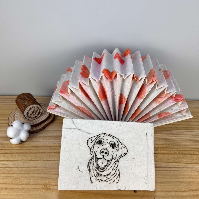 May include: A handmade paper book with a fan-shaped cover featuring orange leaf designs. The book is open to reveal a black and white illustration of a dog with a happy expression.
