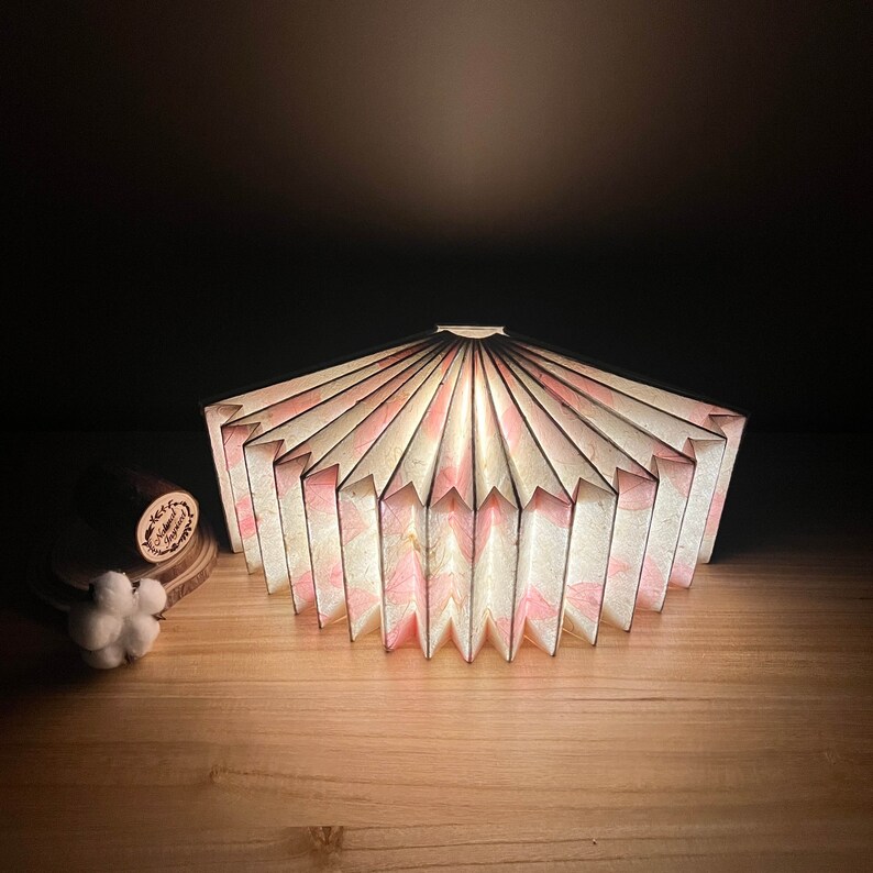 Puede incluir: Una pantalla de l&aacute;mpara de papel blanca y rosa con un dise&ntilde;o plisado. La pantalla est&aacute; encendida y emite un brillo c&aacute;lido.