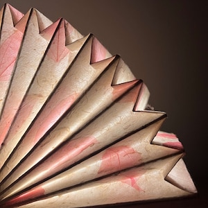 Puede incluir: Un abanico de papel beige y rosa con un dise&ntilde;o delicado y plegado. El abanico est&aacute; hecho de papel fino y transl&uacute;cido con una textura sutil. El abanico est&aacute; plegado en una serie de paneles superpuestos, creando un efecto de capas. El abanico est&aacute; decorado con un patr&oacute;n de hojas rosas.