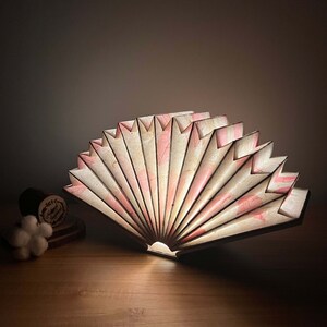 Puede incluir: Una l&aacute;mpara de papel en forma de abanico blanco y rosa con un dise&ntilde;o delicado y plegado. La l&aacute;mpara est&aacute; encendida y emite una luz c&aacute;lida.