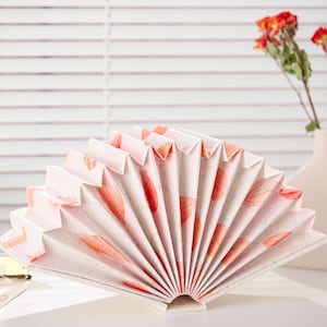 May include: A white paper fan with a pink and orange floral pattern. The fan is folded and standing upright on a white surface.