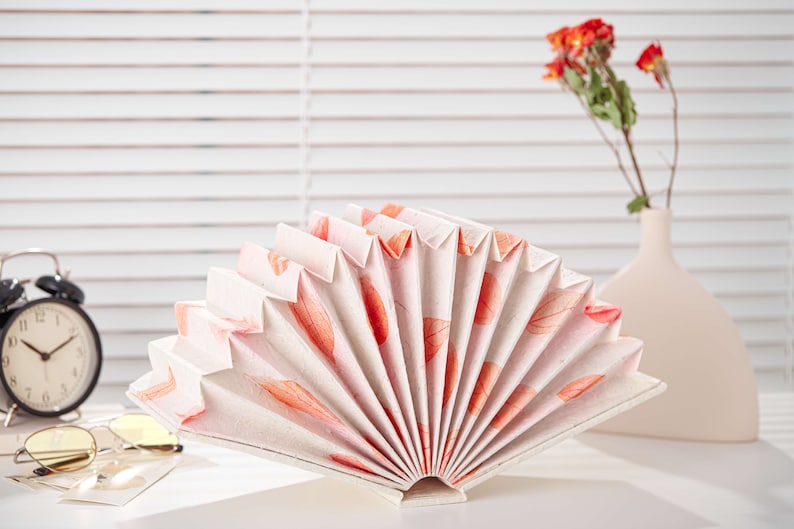 May include: A white paper fan with a pink and orange floral pattern. The fan is folded and standing upright on a white surface.