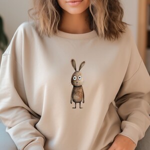 May include: A beige sweatshirt featuring a brown rabbit graphic on the front. The rabbit has large white eyes and a brown body.
