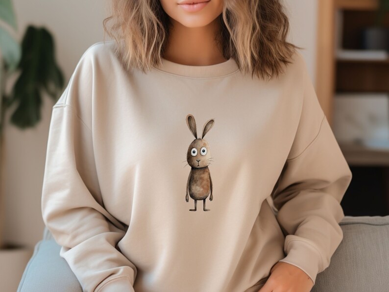 May include: A beige sweatshirt featuring a brown rabbit graphic on the front. The rabbit has large white eyes and a brown body.