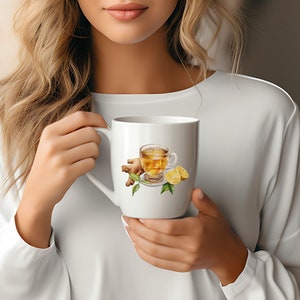 May include: White ceramic mug with a watercolor illustration of a glass of ginger tea with lemon slices. The mug is being held by a person.